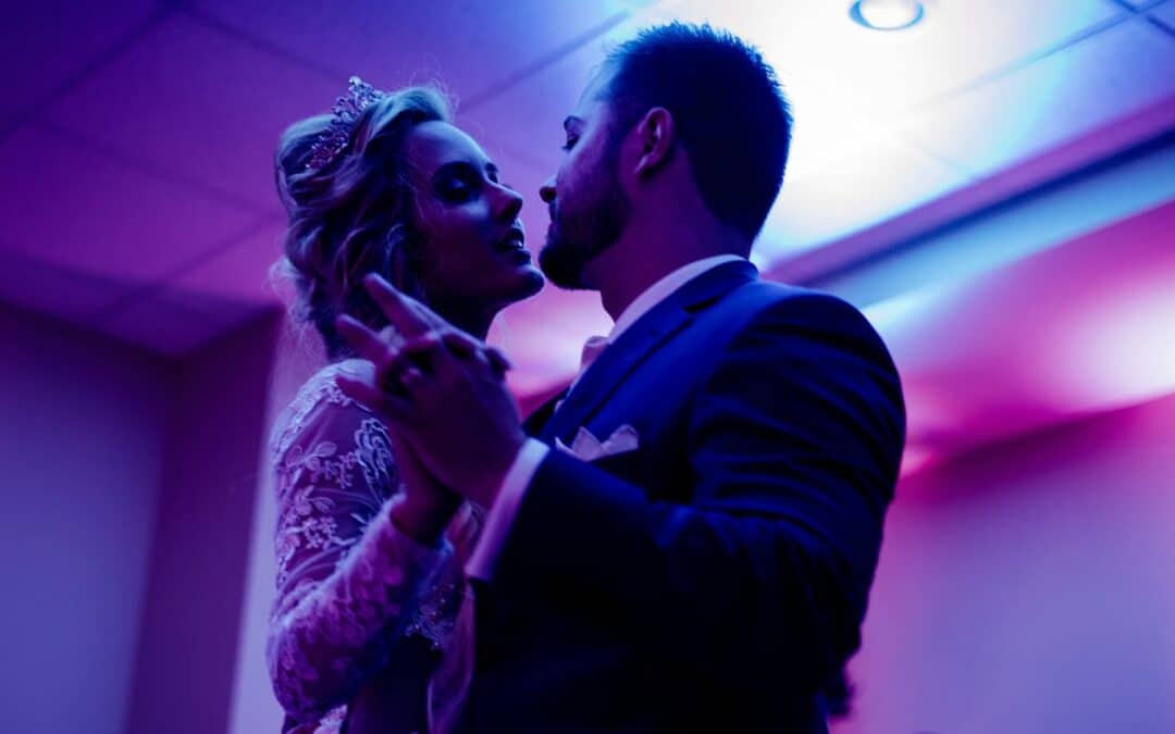 A couple dances intimately under purple lighting, with the woman wearing a lace dress and a tiara, and the man in a suit.