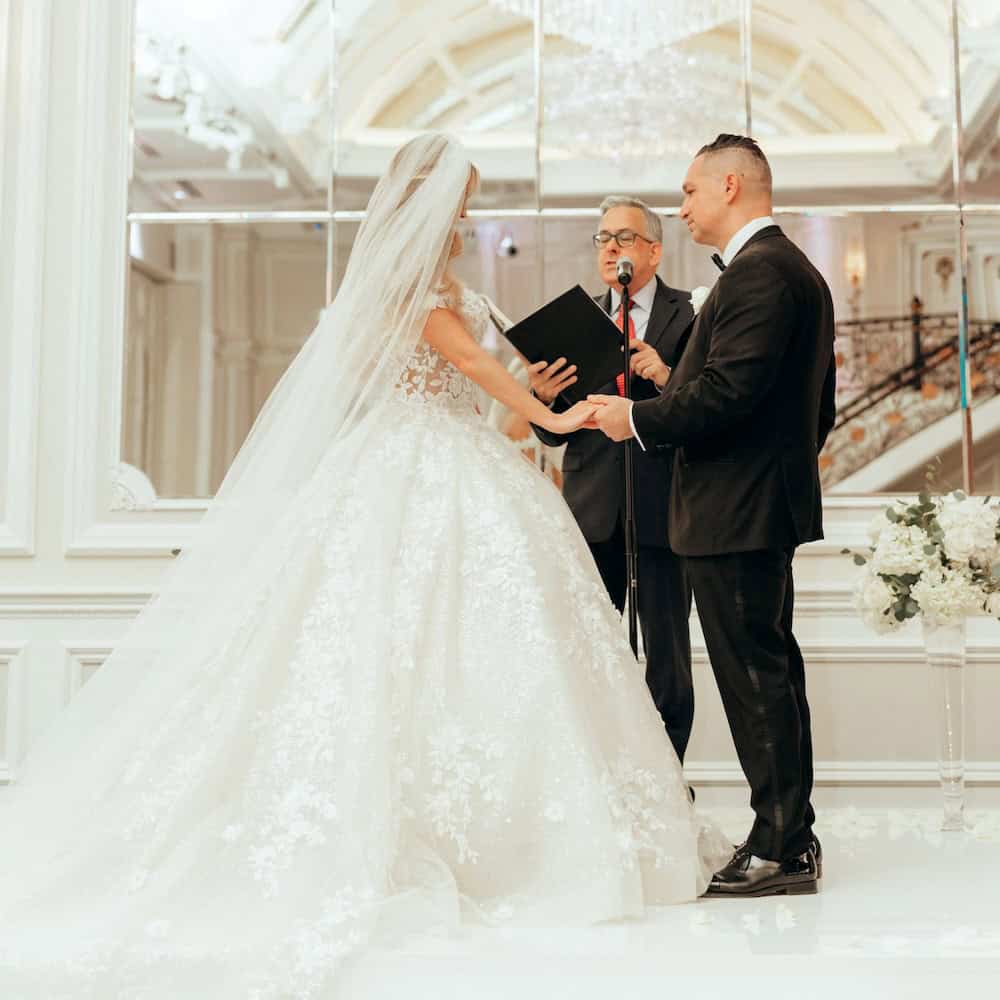 Bride and groom hold hands during their wedding ceremony as an officiant with a book presides. The bride wears a lace gown and veil, the groom a black suit. Elegant venue.