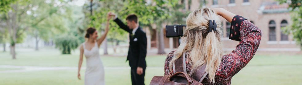 A photographer captures a groom lifting the bride's hand outdoors. They stand on grass, surrounded by trees, with a brick building in the background.